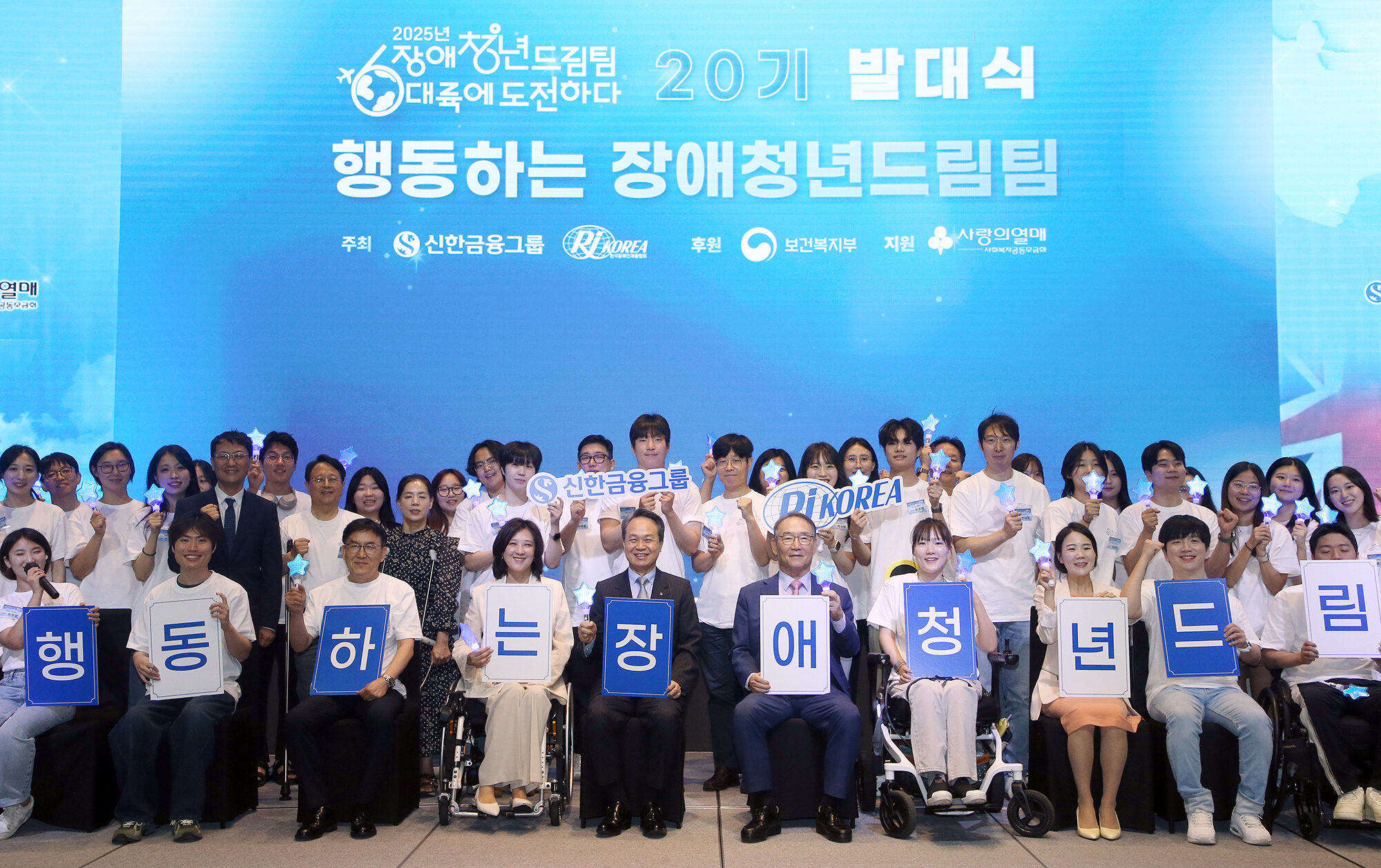 “‘2025 장애청년드림팀, 대륙에 도전하다’ 발대식 현장에서 드림팀 20기 단원들과 관계자들이 무대 앞에서 환하게 웃으며 단체사진을 촬영하고 있다. 앞줄에는 휠체어를 이용하는 청년들도 함께 앉아 있으며, 참가자 전원이 하늘색 배경 앞에서 파란색 별 모양 조명을 들고 활기찬 분위기를 자아낸다. 참가자들은 ‘행동하는 장애청년드림팀’이라는 글자가 적힌 푯말을 들고 있다.”