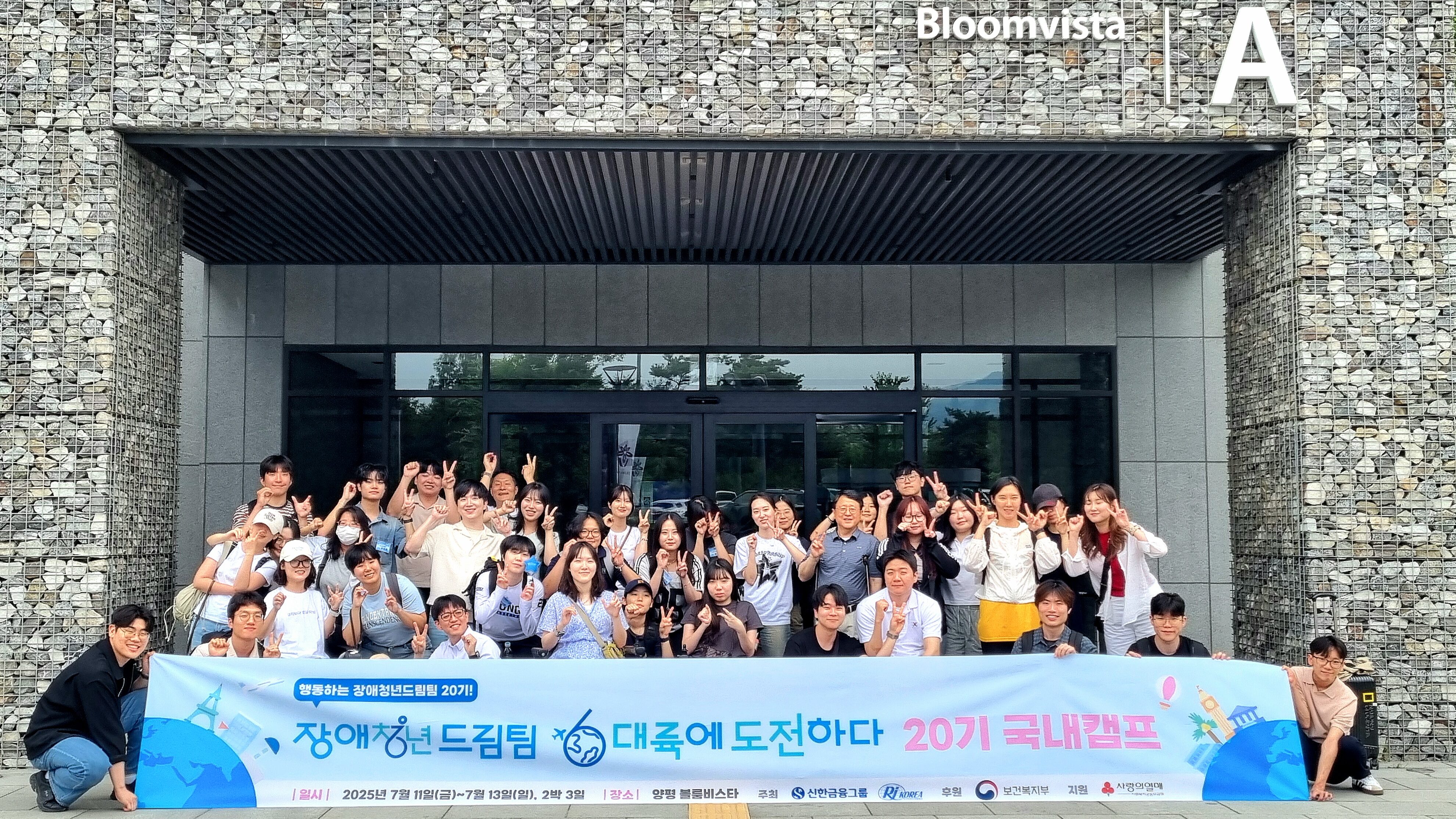 “경기도 양평 블룸비스타 앞에서 열린 ‘2025 장애청년드림팀 20기 국내캠프’ 종료 후, 참가자 40여 명이 건물 A동 앞에 모여 단체사진을 촬영하고 있다. 참가자들은 서로 팔을 두르거나 브이(V) 포즈를 취하며 환하게 웃고 있다. 앞쪽에는 ‘장애청년드림팀, 대륙에 도전하다 – 20기 국내캠프’라는 문구가 적힌 대형 배너가 펼쳐져 있으며, 캠프 일정과 장소, 주최·후원 기관 로고도 포함되어 있다.”
